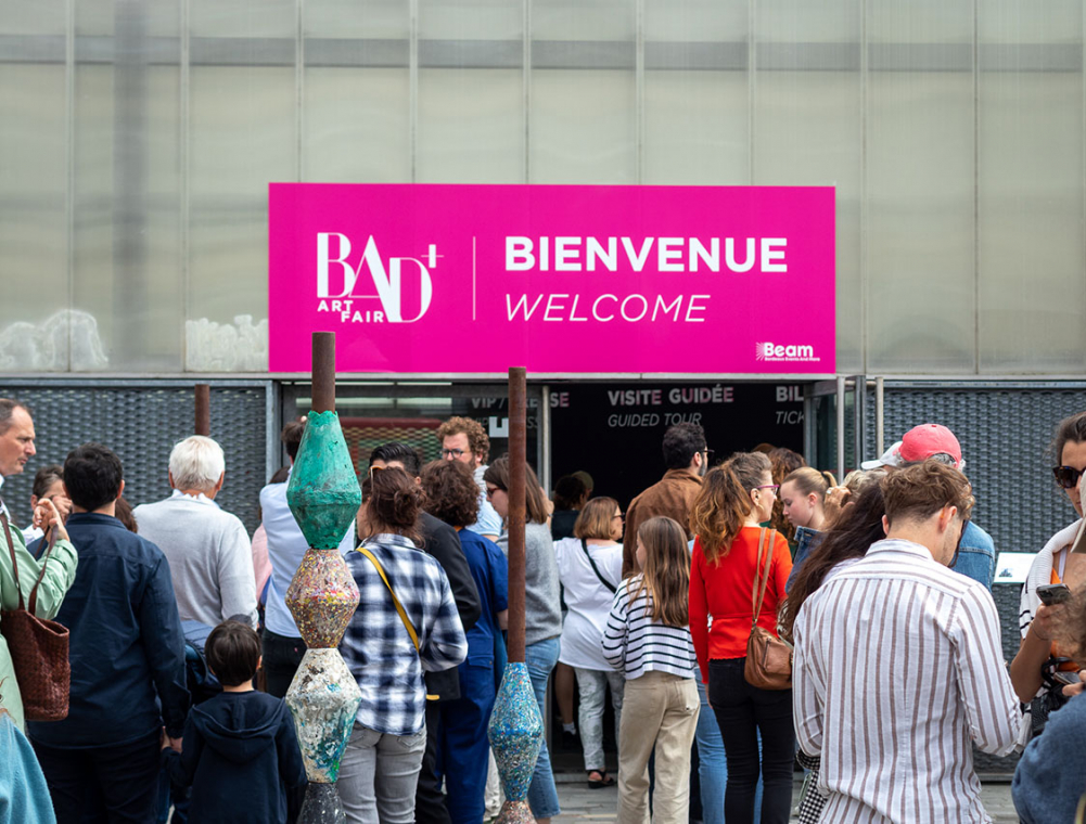 Face à l’entrée du Hangar 14, de nombreuses personnes attendent d’accéder au bâtiment. Une longue file d’attente s’est formée sur le parvis du bâtiment, entre les œuvres exposées à l’occasion de BAD+ International Art Fair.