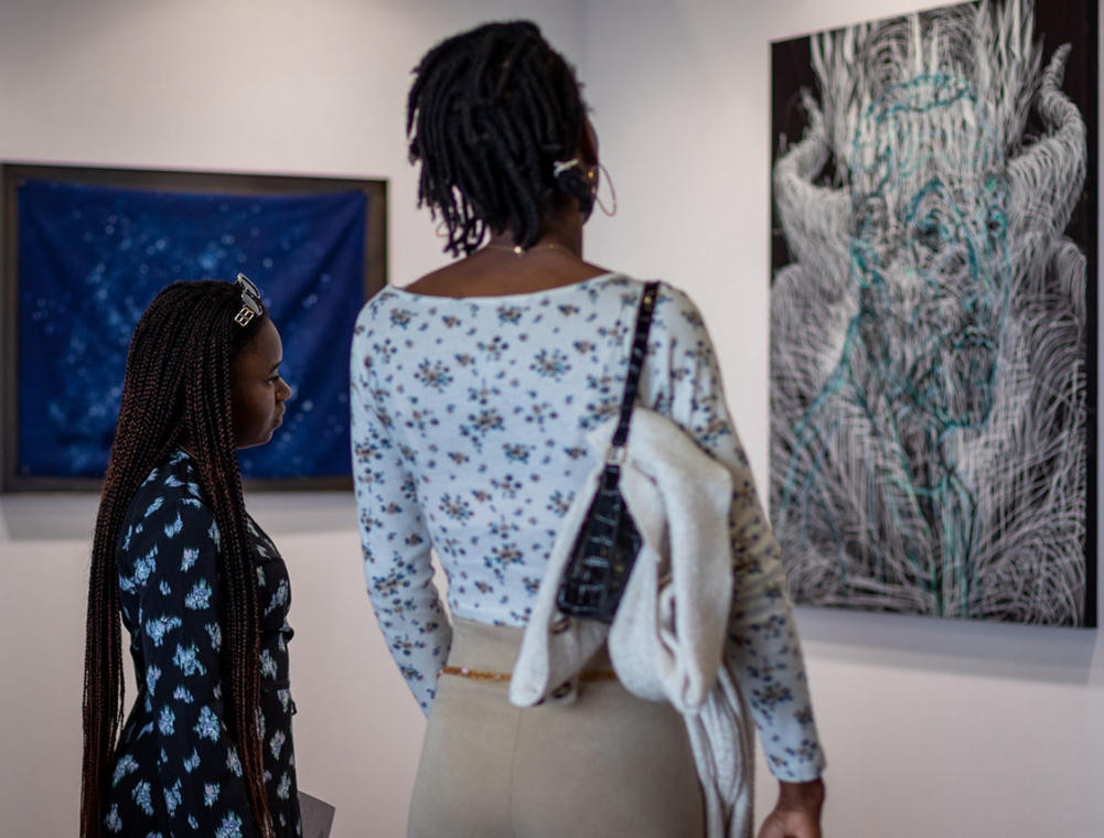 Une adulte et une enfant observent attentivement une œuvre, sur un stand de BAD+ international art fair. L’œuvre, sur fond noir, représente des traits blancs et bleus, tels deux dessins qui se chevauchent.