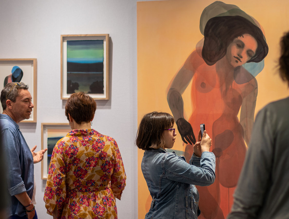 Un homme et une femme discutent devant un triptyque de peinture tandis qu’une femme prend une photo d’une œuvre avec son téléphone.