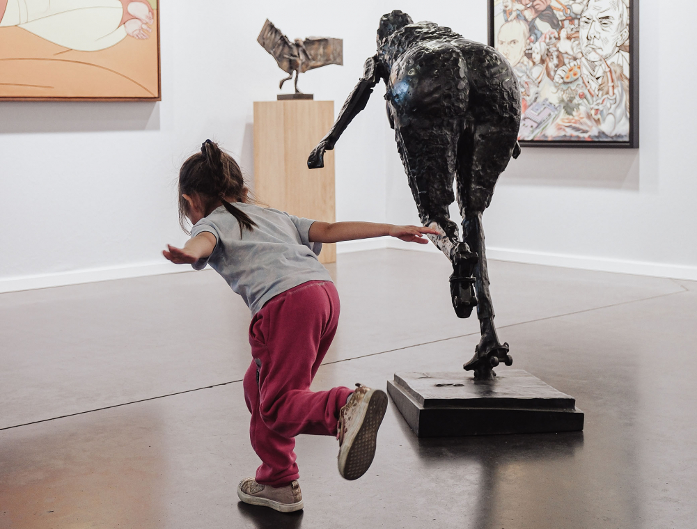 Sur un stand de BAD+ international Art Fair, une statue en bronze présente une personne en patins à roulettes en train de tomber en avant. Sur sa gauche, une petite fille sur un pied reprend la même position, les bras grands ouverts tentant de rattraper son équilibre.