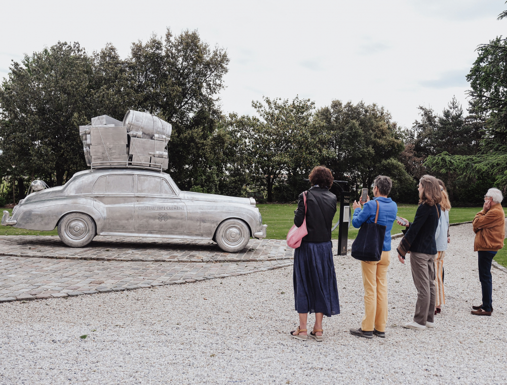 Une sculpture représente une voiture ancienne avec de nombreux bagages sur son toit. Au premier plan plusieurs visiteurs nous tournent le dos, observent la sculpture, l'un d'eux la photographie. En fond, un jardin arboré.