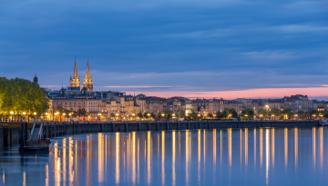 Photo de bâtiments logeant les quais de la Garonne, éclairés par des lumières artificielles. La photo a été prise en soirée, peu avant la nuit et on peut observer le reflet des lumières dans l'eau au premier plan. 