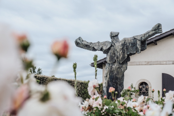 Une grande sculpture grise représentant une silhouette avec les bras ouverts au milieu des fleurs.