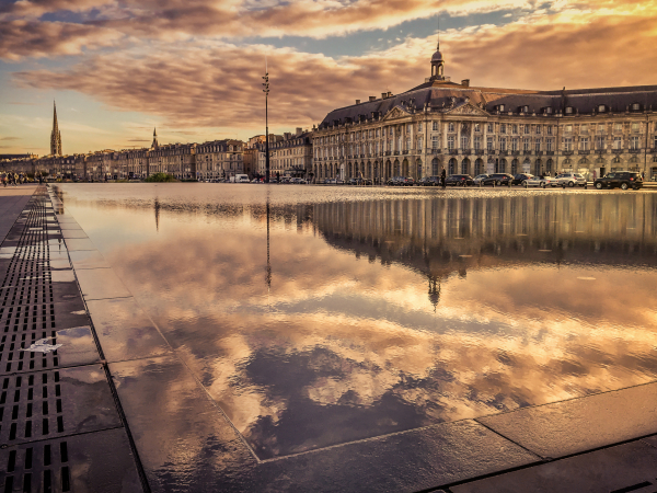 Photo des bâtiments logenant les quais, se refletant dans le miroir d'eau situé place de la Bourse, pendant un coucher de soleil.
