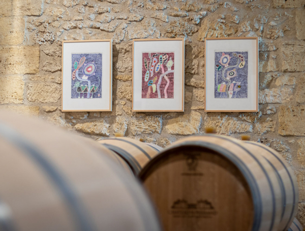 Dans un chai, dont on voit les barriques de vin, trois œuvres colorées sont encadrées sur un mur de pierre.