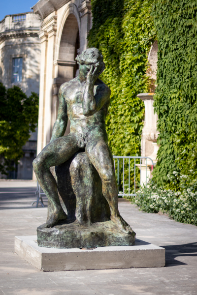 Devant un bâtiment ancien couvert de lierres, une sculpture en bronze représente un homme nu assis sur une pierre. Il se tient la tête dans la main gauche.