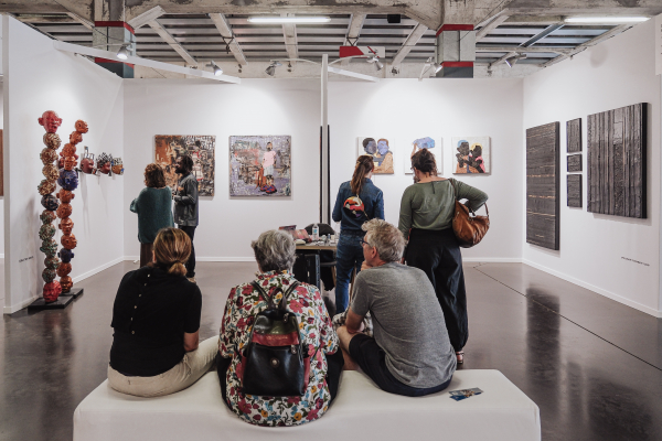 Vue sur un grand stand exposant des peintures et des sculptures. Au premier plan, trois visiteurs assis sur un banc blanc nous tournent le dos. Au second plan, deux groupes de deux visiteurs observent des œuvres de part et d'autre du stand.